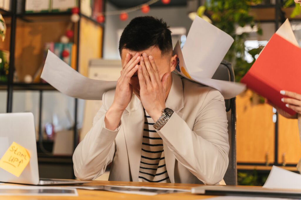 A man is covering his face with papers while sitting at a desk.