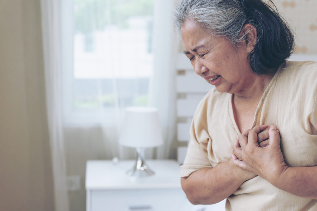 An asian woman is clutching her chest in excruciating pain, showing symptoms of potential atrial fibrillation and stroke.