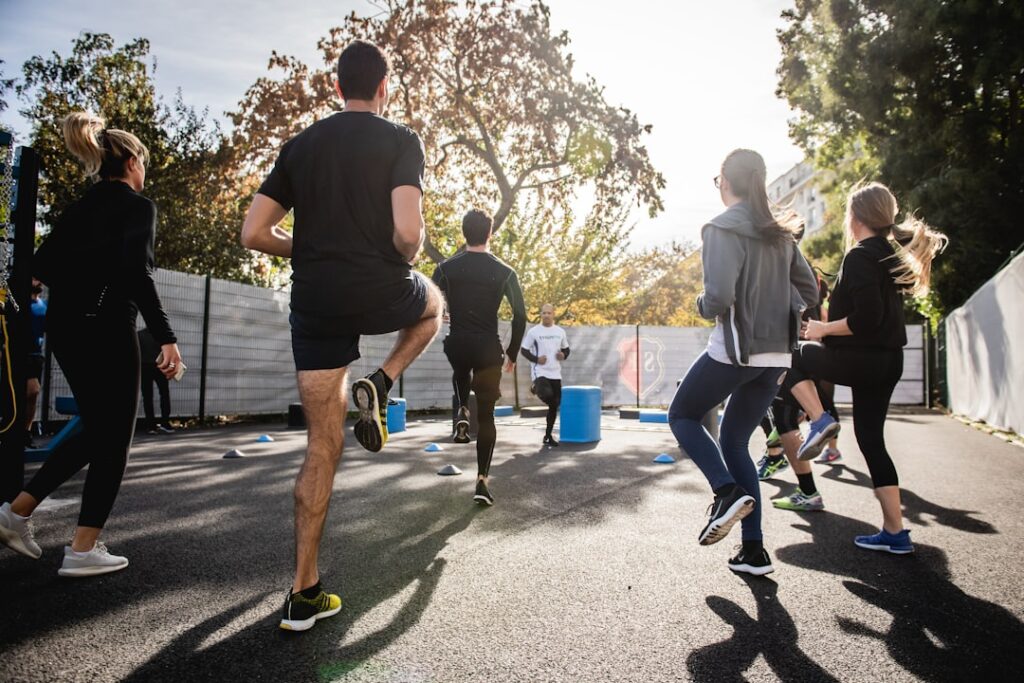 A group of people exercising outdoors on a sunny day. They are performing a workout routine on a paved surface with trees and a fence in the background.