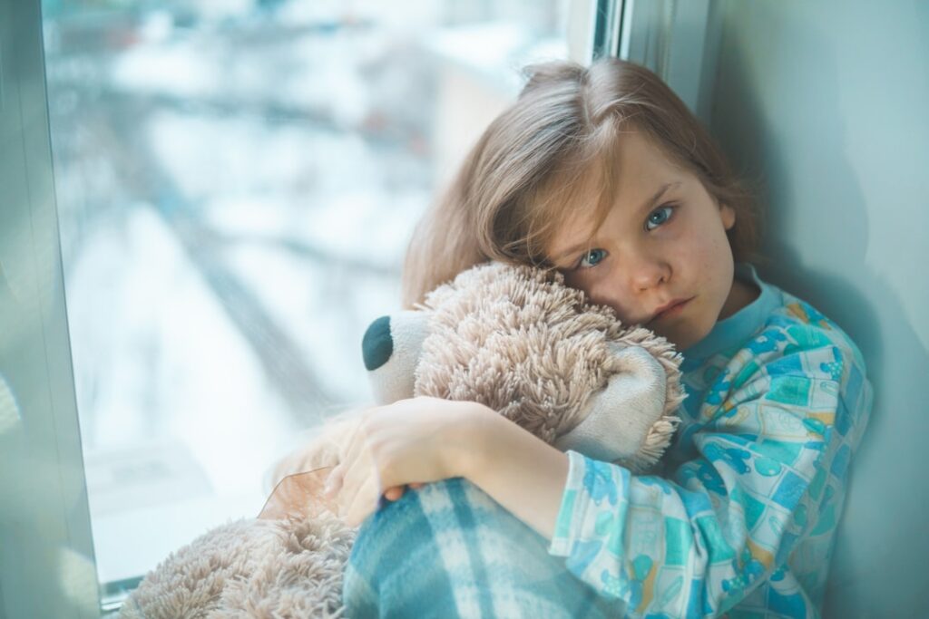 Child in pajamas sitting by a window, holding a teddy bear, gazing outside with a thoughtful expression.