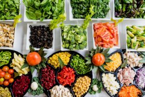 An assortment of fresh vegetables, including spinach, broccoli, tomatoes, olives, corn, peppers, and chickpeas, arranged in bowls and trays on a white surface.