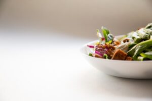A white bowl filled with a fresh salad, including greens, red onion, lentils, and croutons, placed on a white surface.