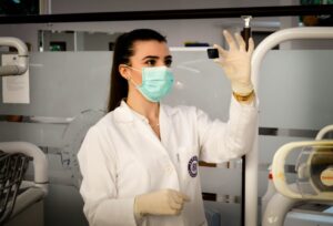 A woman in a lab coat and mask holds up a small black object, examining it. She is in a clinical or laboratory setting.