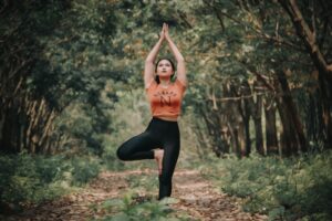 A person practices yoga in the woods, standing in a tree pose, with hands raised overhead and one foot placed on the opposite thigh. Surrounded by trees and fallen leaves.