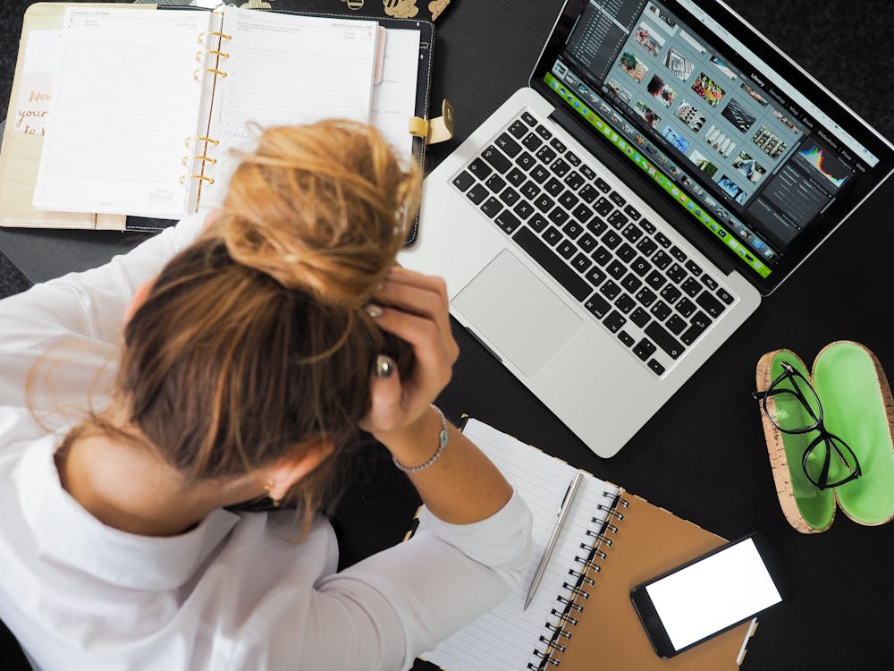 Person at desk with open laptop, notebooks, and smartphone. Hands placed on head, managing stress through focused concentration.